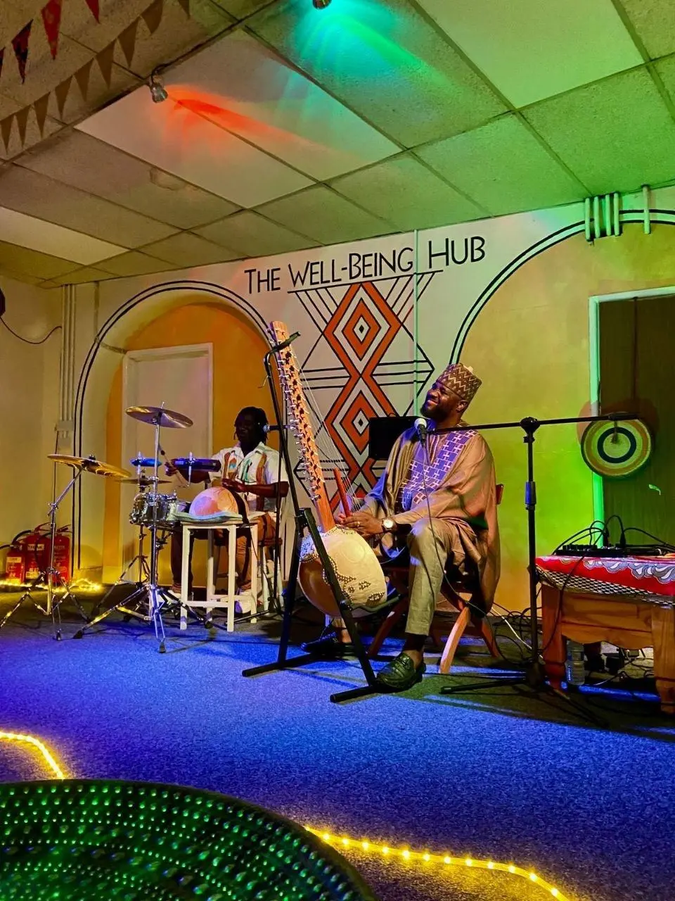 A kora player and a drummer performing under the Wellbeing Hub mural
