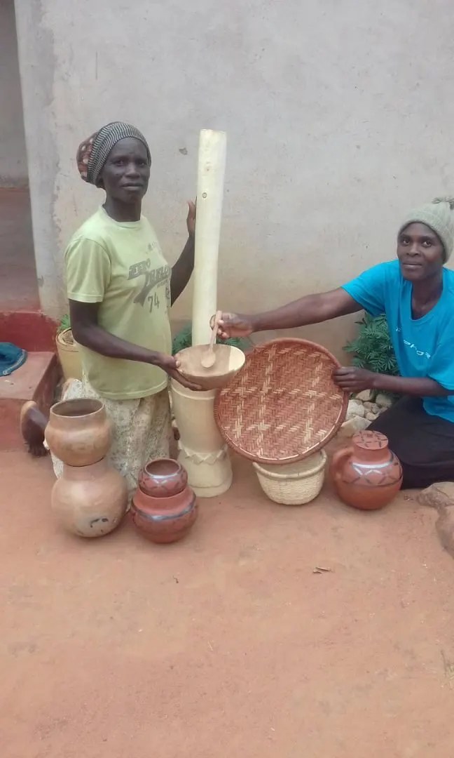 Two women seated beside handmade clay pots, a wooden mortar and woven baskets